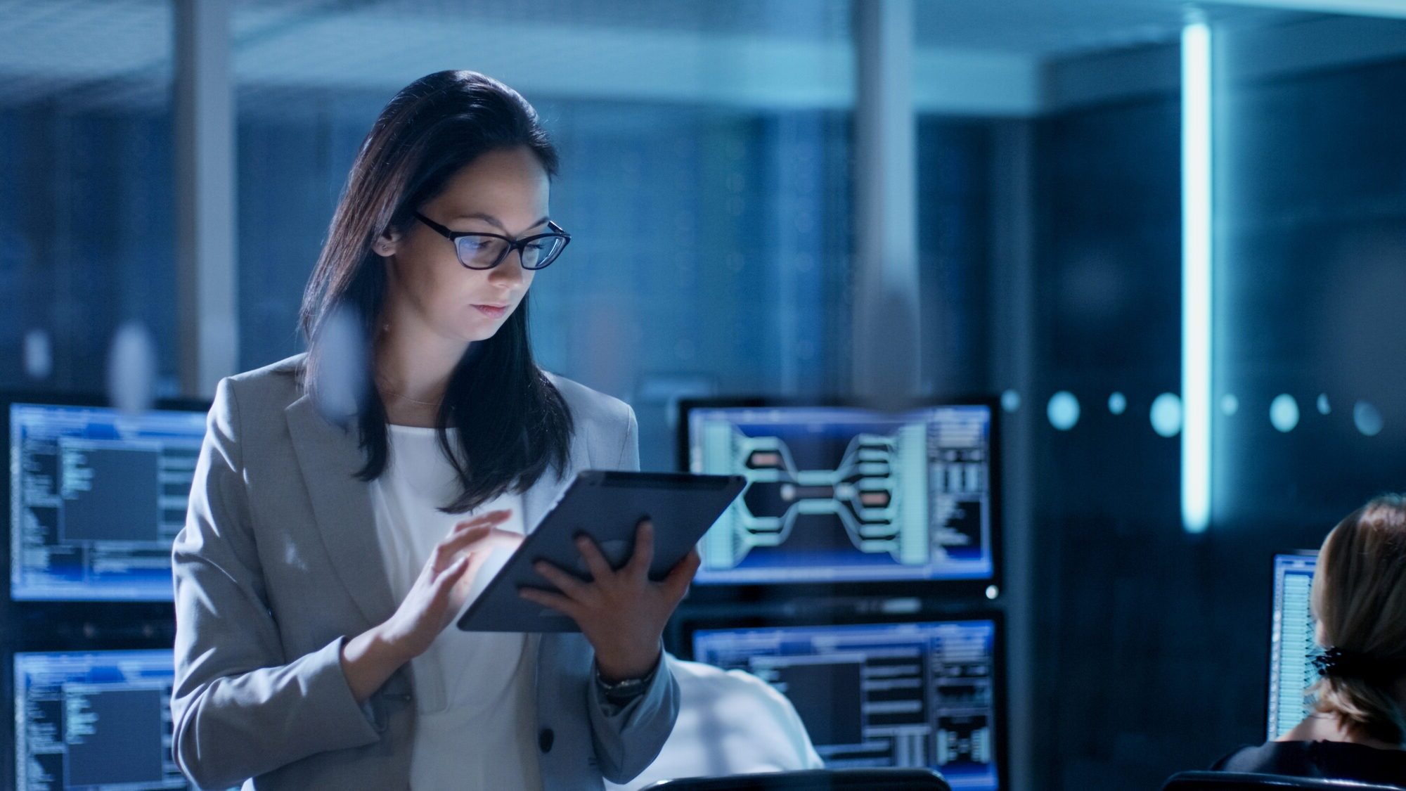 Young Female Government Employee Wearing Glasses Uses Tablet in System Control Center. In the Background Her Coworkers Are at Their Workspaces With Many Displays Showing Valuable Data.